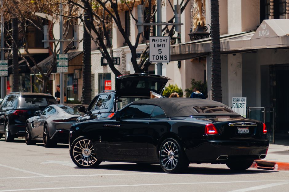 Photograph of a street scene featuring several vehicles, including a black luxury convertible with the top down, parked along the curb in North End. Behind the convertible, a silver SUV with an open trunk is visible, indicating loading or unloading activity. Further down the street, additional cars are parked, with some partially obscured by trees and street signage. The background shows commercial buildings with signage, including a sign that reads 'Vila Blanca' and a prominent weight limit sign indicating a 5-ton restriction. There are pedestrians walking on the sidewalk, and the area appears well-lit with natural daylight. Several palm trees line the street, contributing to a typical urban setting suited for house removals or home relocations, where proper parking and loading logistics are essential. This scene exemplifies the logistics involved in furniture transport and the importance of clear traffic management during moving days, as handled by companies like Man with Van North End.