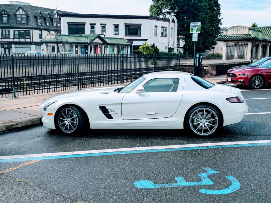 A white Mercedes-Benz SLS AMG sports car with distinctive gullwing doors parked on a street, positioned next to a blue and white marking indicating a designated accessible parking space. The vehicle is adjacent to a curb with an overhanging sidewalk and a black metal fence separating the street from a row of residential buildings with varied architectural styles, including multi-story houses with green and grey roofs. Behind the car, a red vehicle is partially visible, parked parallel to the Mercedes-Benz. The scene is illuminated by daylight with light cloud cover, and the street surface appears slightly wet. This image captures the process of parking in urban areas, which can be relevant to house removals and vehicle transport logistics with companies like Man with Van North End, especially for home relocation or furniture transport during moving days.
