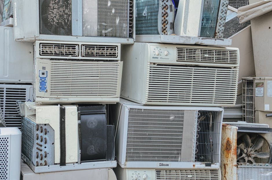 A collection of old, discarded window air conditioning units stacked outdoors on a concrete surface, with some showing rust and damage. The units vary in size and design, with metal grilles, control panels, and vents visible. Several units are partially covered in dust and dirt, indicating they have been unused for some time. In the background, there are additional appliances and containers, suggesting an area used for bulky waste disposal. The environment appears to be an outdoor storage or collection site, relevant for house removals and disposal tips, with a focus on the proper handling and disposal of large appliances, as seen with the company Man with Van North End.