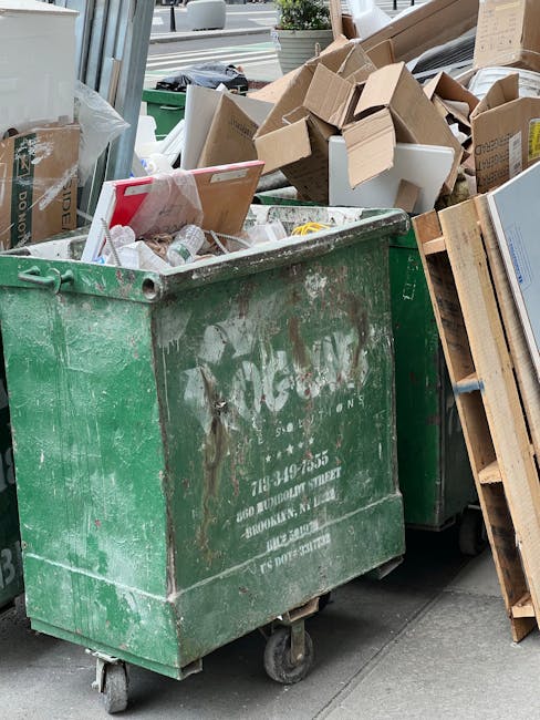 A large green waste disposal container with visible wear and rust, positioned outdoors on a paved surface, filled with assorted household rubbish including cardboard boxes, plastic bottles, and paper. Beside the container are additional cardboard boxes and packaging materials, some partially collapsed or damaged, indicating recent collection or disposal activity. The surrounding environment includes a glass window or door reflecting the street scene, with glimpses of nearby buildings and outdoor furniture. The scene depicts a typical loading area used in house removals or home relocation processes, where bulk waste such as furniture, appliances, and packaging materials are prepared for disposal or recycling, consistent with professional removal services like those provided by Man with Van North End.