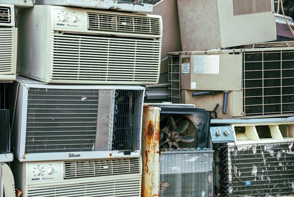 A collection of old, discarded window air conditioning units stacked outdoors on a concrete surface, with some showing rust and damage. The units vary in size and design, with metal grilles, control panels, and vents visible. Several units are partially covered in dust and dirt, indicating they have been unused for some time. In the background, there are additional appliances and containers, suggesting an area used for bulky waste disposal. The environment appears to be an outdoor storage or collection site, relevant for house removals and disposal tips, with a focus on the proper handling and disposal of large appliances, as seen with the company Man with Van North End.