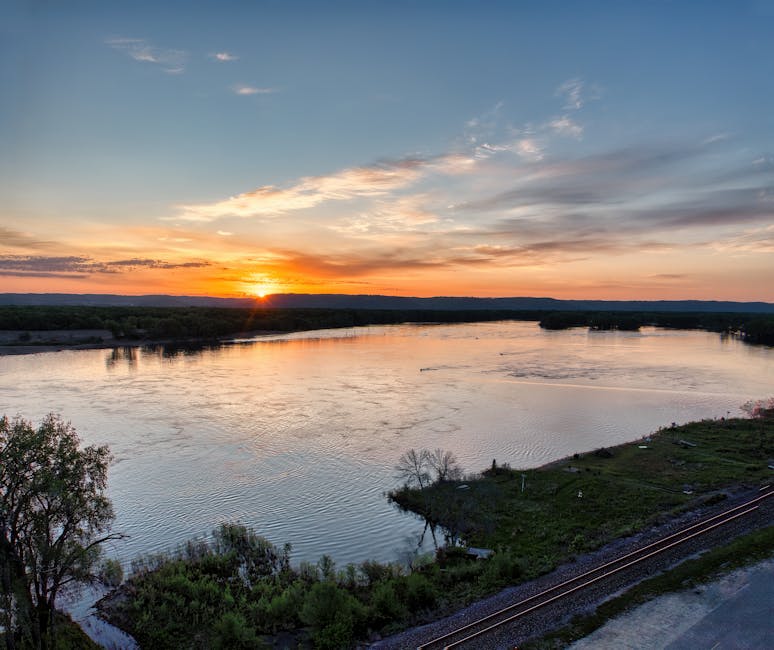 A wide view of a river scene at sunset with the sky displaying orange and yellow hues blending into blue and gray clouds. The river’s surface reflects the sky’s colors, and gentle ripples are visible along its length. Along the riverbank, there are patches of grass, small trees, and shrubs, with a railway track running parallel to the water on the right side of the image. In the foreground, trees and vegetation frame the scene, and the overall landscape appears calm and natural, providing a peaceful outdoor setting during the late evening.