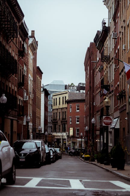 A narrow urban street scene in North End with tall, multi-storey brick buildings on both sides, featuring fire escape balconies and small decorative windows. Parked cars line both sides of the street, some partially on the pavement. In the foreground, a section of the road displays white crosswalk markings and a speed restriction sign on the right side. The street appears to be a typical residential or mixed-use area with narrow sidewalks, planters, and lighting fixtures attached to building facades. The sky above is overcast, casting diffuse lighting on the scene. This setting provides context for house removals or furniture transport, with the environment suitable for loading or unloading for moving services, as occasionally handled by companies like Man with Van North End.