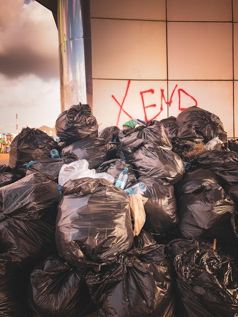 A large pile of black garbage bags filled with waste materials, including visible plastic bottles, is stacked outside a modern building with a reflective metal column and tiled wall. The wall has graffiti with the word 'XEND' spray-painted in red. The scene appears to be part of a waste disposal or clearance process associated with house removals or relocation services, with the bags likely containing household items or rubbish awaiting collection. The background shows a cloudy sky and distant cityscape, indicating an urban environment. Man with Van North End may be involved in transporting or disposing of such waste during house moving or clearance operations.