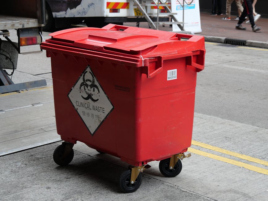 A red clinical waste disposal bin with a hinged lid and four casters situated on a concrete pavement. The bin has a biohazard symbol and a 'Clinical Waste' label on its side, indicating its use for hazardous medical refuse. Behind the bin, a van with a partially open rear door and a metal ramp is visible, suggesting loading or unloading activities related to house removals or waste disposal. In the background, there are pedestrians walking on the street and some street markings, with the scene illuminated by natural daylight. The image depicts the process of handling waste materials within a home relocation or clearance context, consistent with professional moving and waste management services offered by Man with Van North End.