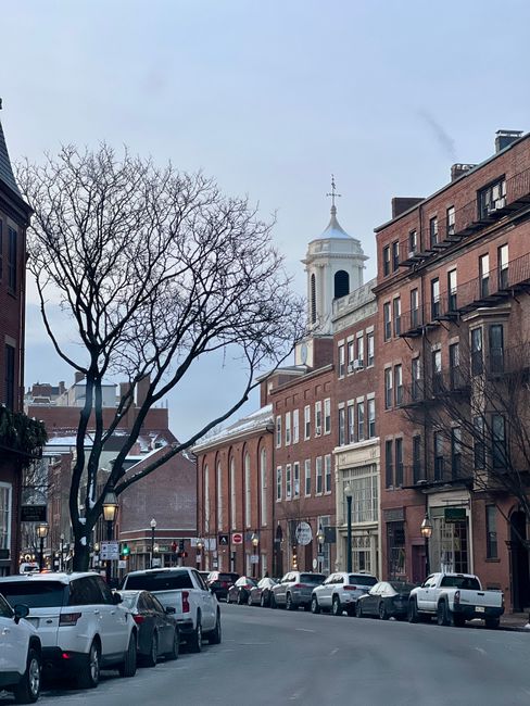 A street scene in North End showing a row of parked cars along the pavement, with a mixture of modern and historic brick buildings lining the street. In the background, a white church with a tall clock tower and spire is visible against a cloudy sky. A leafless tree with bare branches stands to the left, and street lamps are illuminated along the sidewalk. The overall scene reflects a quiet residential area suitable for home relocation or furniture transport. Man with Van North End provides removals and moving services that typically involve loading and unloading household items in such urban environments, often requiring city council permits for parking or loading zones as indicated by the page title.