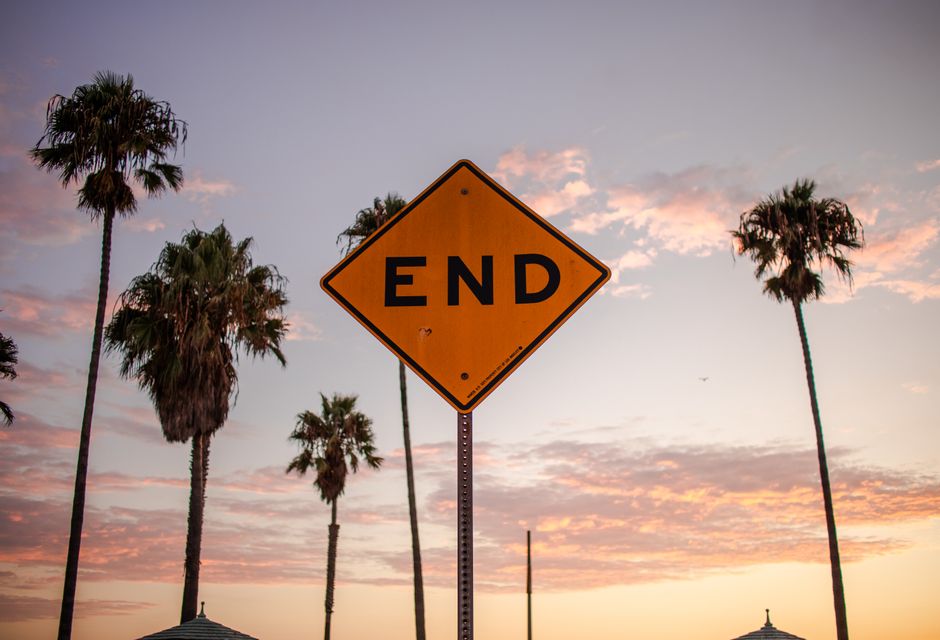 A close-up view of a yellow diamond-shaped road sign with the word 'END' printed in bold black letters, mounted on a metal pole, set against a backdrop of a sunset sky with scattered pink and orange clouds. Several tall palm trees with slender trunks and fan-shaped fronds are visible behind the sign, creating a tropical outdoor scene. The scene is outdoors, likely in a warm climate area, with natural lighting that highlights the sign and the trees. The environment appears calm, and the sign is positioned at the edge of a roadway or property boundary, indicating the conclusion of a particular zone or pathway. This image supports content related to house removals and relocation services, emphasizing the importance of proper planning and adherence to local regulations during home relocation or furniture transport, with [COMPANY_NAME] providing moving and removal assistance in the NORTH END area.