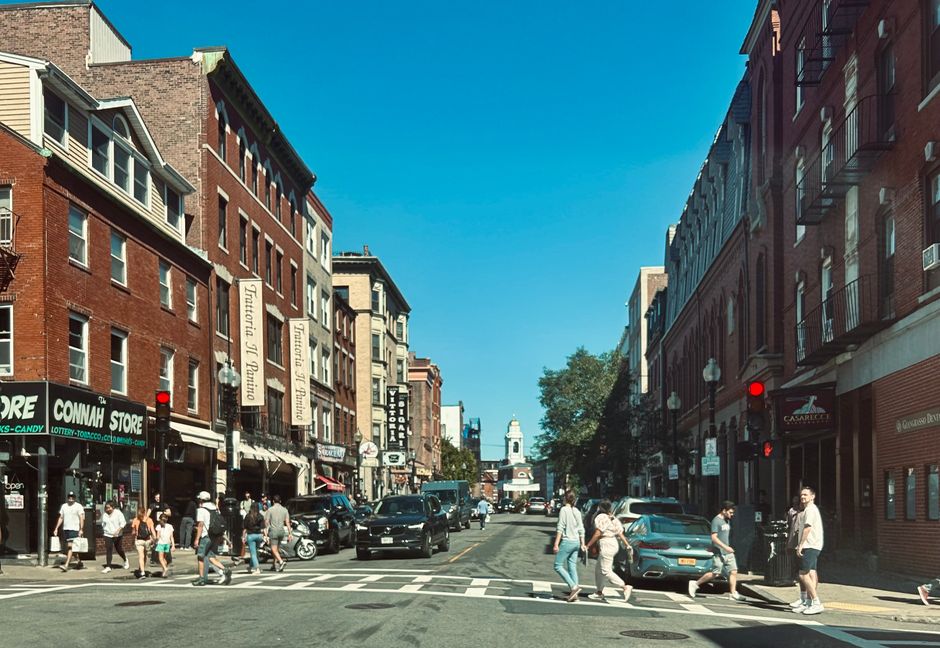 A street scene in North End showing a row of parked cars along the pavement, with a mixture of modern and historic brick buildings lining the street. In the background, a white church with a tall clock tower and spire is visible against a cloudy sky. A leafless tree with bare branches stands to the left, and street lamps are illuminated along the sidewalk. The overall scene reflects a quiet residential area suitable for home relocation or furniture transport. Man with Van North End provides removals and moving services that typically involve loading and unloading household items in such urban environments, often requiring city council permits for parking or loading zones as indicated by the page title.