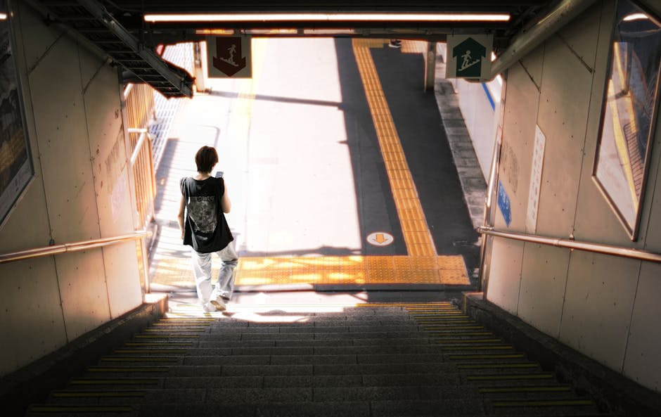 A person with short dark hair, dressed in a black sleeveless top and light-colored pants, is descending a concrete staircase at a transportation station. They are carrying an item in their right hand and have a black shoulder bag hanging across their body. The staircase is enclosed by metal railings painted in a copper colour, with yellow tactile paving on the steps for safety. The station is illuminated by natural sunlight pouring in from the entrance at the bottom, which is surrounded by signage including emergency exit symbols and directional signs. Outside the station, the pavement features a tactile guide path with yellow textured strips, indicating a pedestrian pathway. This scene depicts a typical urban setting supporting home relocation or moving logistics, with clear signage and accessible features. Man with Van North End occasionally offers house removals and furniture transport services, focusing on loading and unloading processes in such environments.