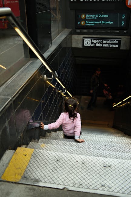 A young girl, wearing a pink and white striped sweater with white cuffs, is sitting on a metal staircase inside a building near North End station. She is facing away from the camera, with her right hand resting on the black tiled wall beside her. The staircase has metallic steps with a textured surface for grip, and yellow warning tape marks the edge of each step. A metal handrail runs along the left side, illuminated by overhead lighting. In the background, there are several people walking down the stairs, with a digital sign displaying train destinations such as Uptown & Queens and Downtown & Brooklyn, as well as an information sign indicating an agent is available at the entrance. The environment appears to be a station corridor, with a dimly lit, utilitarian atmosphere typical of urban transit facilities. This scene may relate to home relocation or moving logistics, as part of a household move within the station area, handled by Man with Van North End's removals service.