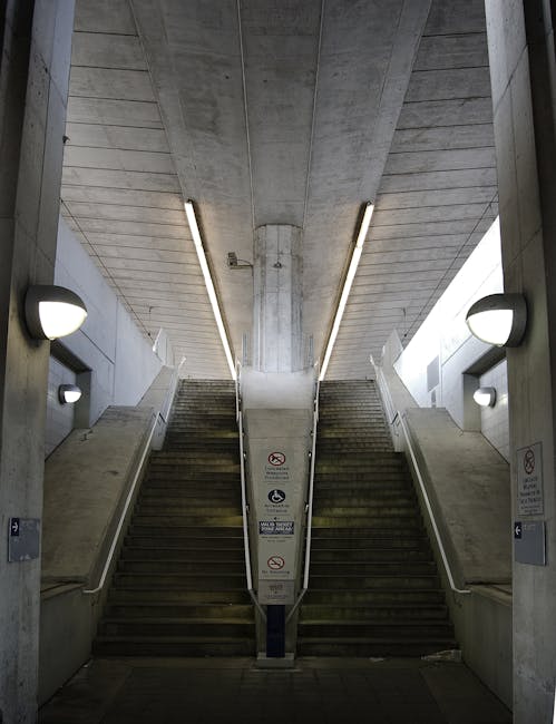 A person with short dark hair, dressed in a black sleeveless top and light-colored pants, is descending a concrete staircase at a transportation station. They are carrying an item in their right hand and have a black shoulder bag hanging across their body. The staircase is enclosed by metal railings painted in a copper colour, with yellow tactile paving on the steps for safety. The station is illuminated by natural sunlight pouring in from the entrance at the bottom, which is surrounded by signage including emergency exit symbols and directional signs. Outside the station, the pavement features a tactile guide path with yellow textured strips, indicating a pedestrian pathway. This scene depicts a typical urban setting supporting home relocation or moving logistics, with clear signage and accessible features. Man with Van North End occasionally offers house removals and furniture transport services, focusing on loading and unloading processes in such environments.