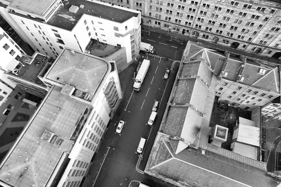 An aerial view taken from a high vantage point showing a narrow urban street lined with multi-storey residential and commercial buildings. Several parked cars, including small passenger vehicles, are visible along the street and in parking areas adjacent to the buildings. The street is partly occupied by a white moving truck with a closed cargo area positioned for loading or unloading, and a man from Man with Van North End is seen carrying a large cardboard box towards the building's entrance. The buildings display flat and pitched roofs, with visible features such as vents, chimneys, and rooftop equipment. Packaging materials like cardboard boxes, plastic wrap, and packing blankets are present both outside and near the entrance. A rolling trolley and straps are also visible, indicating a home relocation or furniture transport process as part of a professional removals service. The scene captures the typical logistics and equipment involved in house removals within an urban environment, highlighting the company's mass city-wide moving operations, all under natural daylight with clear visibility.