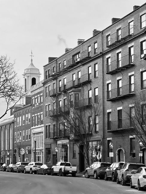 A black-and-white image of a city street scene captured during daytime, showing a row of parked cars lining the cobbled pavement on both sides of the street. Multi-storey buildings with storefronts an