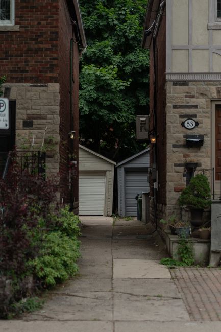 The image shows a narrow alleyway between two residential buildings, leading to a small parking area with two white single-door garages positioned side by side at the end of the alley. The alley is paved with concrete and bordered by plants and shrubs, with a large leafy tree visible in the background. On the right side, a house number 53 is mounted on the wall above a black mailbox, and potted plants and garden ornaments are arranged on the doorstep. The left side features a brick wall and garden greenery. The scene is well-lit with natural daylight, and the setting suggests a typical urban or suburban residential environment suitable for house removals or furniture transport. This photograph, taken by Man with Van North End, demonstrates the type of narrow passageways that may require careful planning during home relocation or moving services to navigate furniture and boxes through tight spaces effectively.