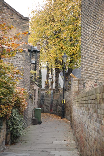 The image shows a narrow alleyway between two residential buildings, leading to a small parking area with two white single-door garages positioned side by side at the end of the alley. The alley is paved with concrete and bordered by plants and shrubs, with a large leafy tree visible in the background. On the right side, a house number 53 is mounted on the wall above a black mailbox, and potted plants and garden ornaments are arranged on the doorstep. The left side features a brick wall and garden greenery. The scene is well-lit with natural daylight, and the setting suggests a typical urban or suburban residential environment suitable for house removals or furniture transport. This photograph, taken by Man with Van North End, demonstrates the type of narrow passageways that may require careful planning during home relocation or moving services to navigate furniture and boxes through tight spaces effectively.