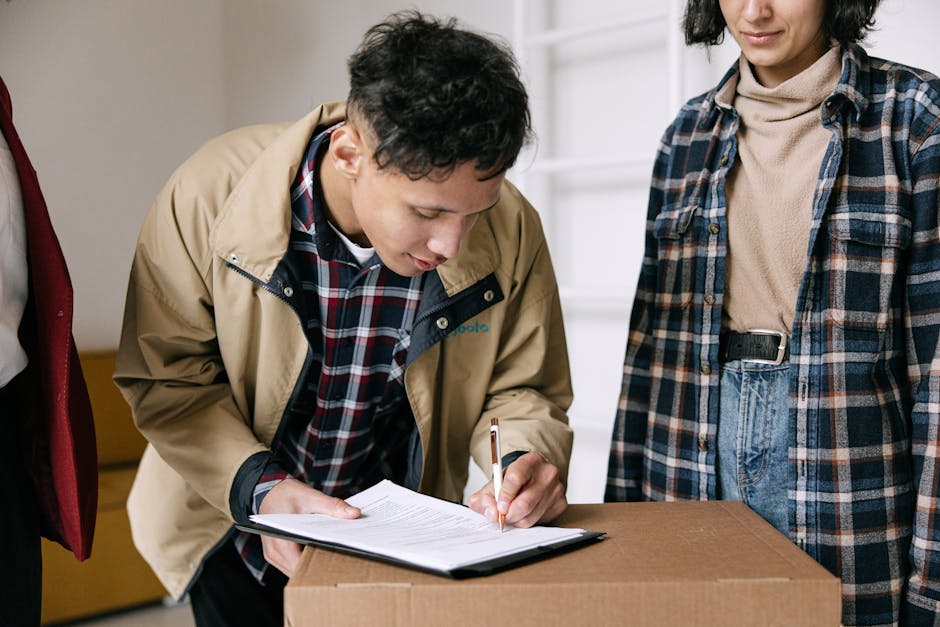A man with short dark hair, wearing a beige jacket over a checked shirt, is inside a property and is signing a document on a clipboard placed on top of a cardboard box. Next to him, a woman with dark hair tied back, wearing a beige turtleneck and a blue plaid shirt, stands observing. The scene appears to be part of a home relocation or packing process, with the individuals engaged in paperwork related to furniture transport or moving logistics. The background includes a white door and shelving, indicating an indoor setting, with natural lighting enhancing the clarity of the objects and individuals involved in the moving activity. Man with Van North End is performing or overseeing a loading or packing stage, supporting home removals through organized documentation of household items prepared for transport.