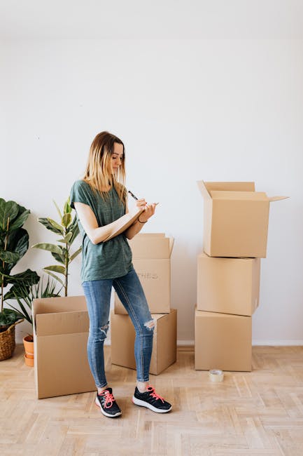 A young woman with shoulder-length blonde hair, dressed in a green T-shirt, ripped blue jeans, and black sneakers with pink accents, standing indoors on a light wood floor next to several brown cardboard moving boxes. She is holding a pen and a small notebook, appearing to take notes or check items during a home relocation or packing process. Behind her, there is a large green houseplant with broad leaves in a woven basket, and the plain white wall suggests a clean, organized environment. To her right, there is an open top cardboard box with its flaps folded outward, indicating packing or unpacking activity as part of furniture transport or move preparation, consistent with a professional removals service such as Man with Van North End.