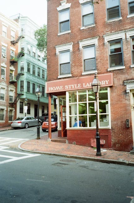 The image shows a multi-storey brick building with a ground-floor storefront labeled 'Home Style Laundry' featuring large glass windows and a red brick exterior. In front of the shop, there is a small paved sidewalk area with a decorative black lamppost and a black bollard. Parked cars are visible along the street, including a grey vehicle and a red vehicle, indicating a typical urban neighbourhood scene. The street is cornered with a pedestrian crossing visible at the intersection. The building's upper floors have multiple windows, some with white trim, and the background includes other older buildings with various architectural styles, suggesting an area where professional house removals and furniture transport could take place. The lighting is natural daylight, highlighting the urban environment conducive to home relocations with careful loading and unloading of furniture and moving boxes by [COMPANY_NAME].