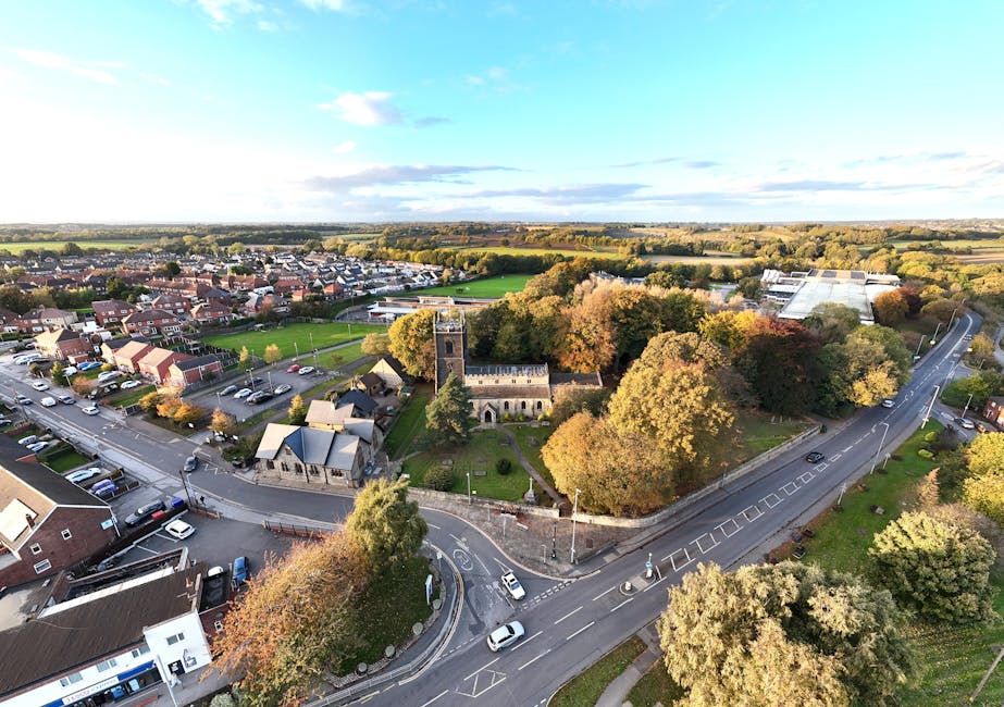 Aerial view of North End showing a small historic church with a square tower and stone walls, surrounded by a grassy churchyard with mature trees displaying autumn foliage. The church is located at a T-junction where a main road curves past it, with several cars parked along the street and a few vehicles in motion. Adjacent to the church, there are residential buildings including semi-detached and terraced houses, many with small front gardens, and some with pitched roofs. To the right, part of a large industrial or warehouse building with a flat roof is visible, along with a sports field and open green spaces further in the distance. The overall scene includes overcast sky with some patches of blue, and the area appears to be part of a suburban neighbourhood with a mix of greenery and built structures, ideal for local house removals or furniture transport as handled by Man with Van North End, especially during a home relocation or moving process involving packing and loading.