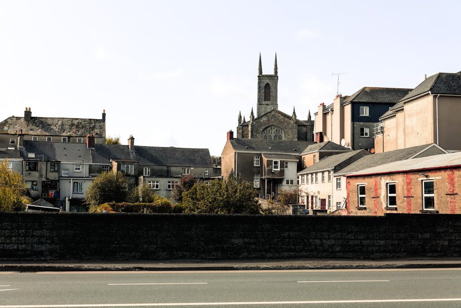 A street view in North End showing a mix of residential buildings and a prominent church with a tall, pointed spire. The houses include terraced and semi-detached styles, with various exterior materials such as brick and weatherboard, and are situated behind a low stone wall along the pavement. Some buildings have visible windows, roofs with tiles, and residential features like chimneys and satellite dishes. The scene is captured during daylight with clear skies, indicating a typical urban environment. This setting is relevant to house removals and furniture transport, illustrating the typical architecture and environment encountered during relocation or moving services, as provided by Man with Van North End.